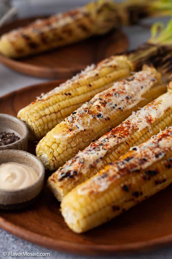 4 ears of Grilled Mexican Street Corn on the Cob on brown plate with 2 dipping sauces and a grilled corn on the cob in the background.
