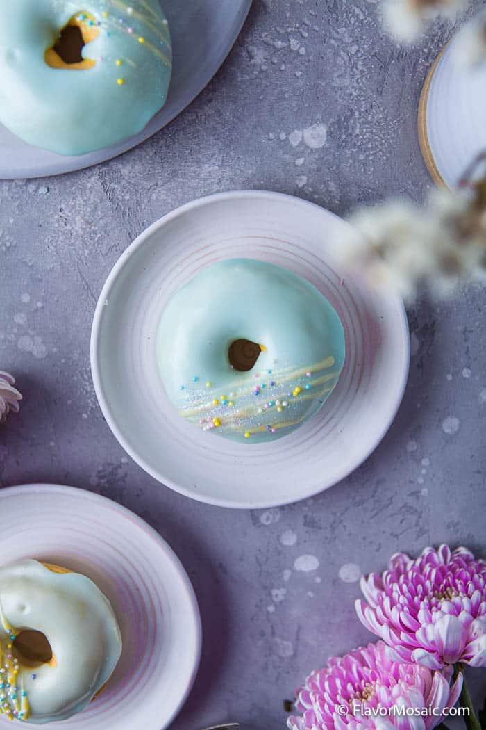 Overhead view of 3 individual plates, each with a Vanilla Frosted Donuts with teal-blue frosting and yellow glitter with a partial view of a pink flower in the bottom corner and a blurred white flower and partial view of a white plate in the top right corner.