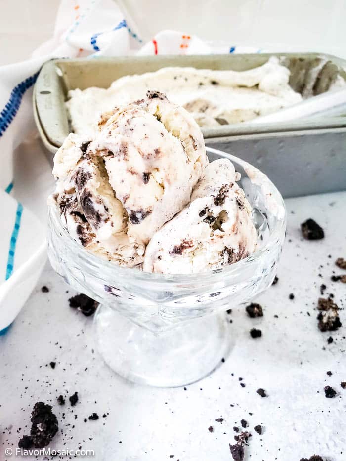 2 scoops of Cookies and Cream Ice Cream in a small glass ice cream dish in front of a loaf pan with homemade Cookies and Cream No Churn Ice Cream and surrounded by crumbled pieces of Oreo Cookes.
