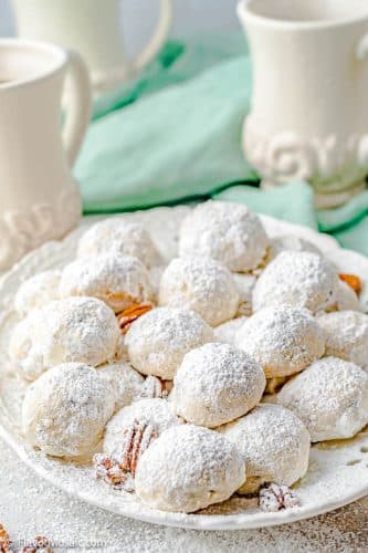 plate with about a dozen Mexican Wedding Cookies covered in powdered sugar and with a few pecan halves sprinkled on the plate, with 3 white vases and a teal napkin in the background.