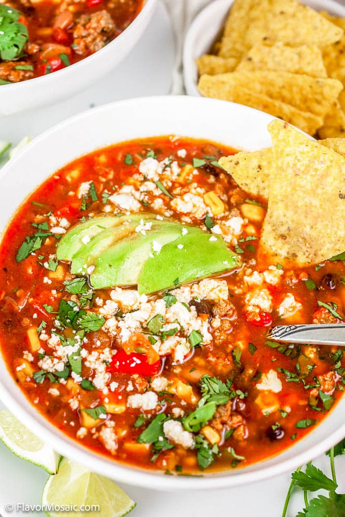 Overhead view of white bowl with Turkey Taco Soup with cheese crumbles, chopped cilantro, avocado slices and tortilla chips, and the bowl is surrounded by limes, tortilla chips, cilantro, and a partial view of a second bowl in the upper left corner.