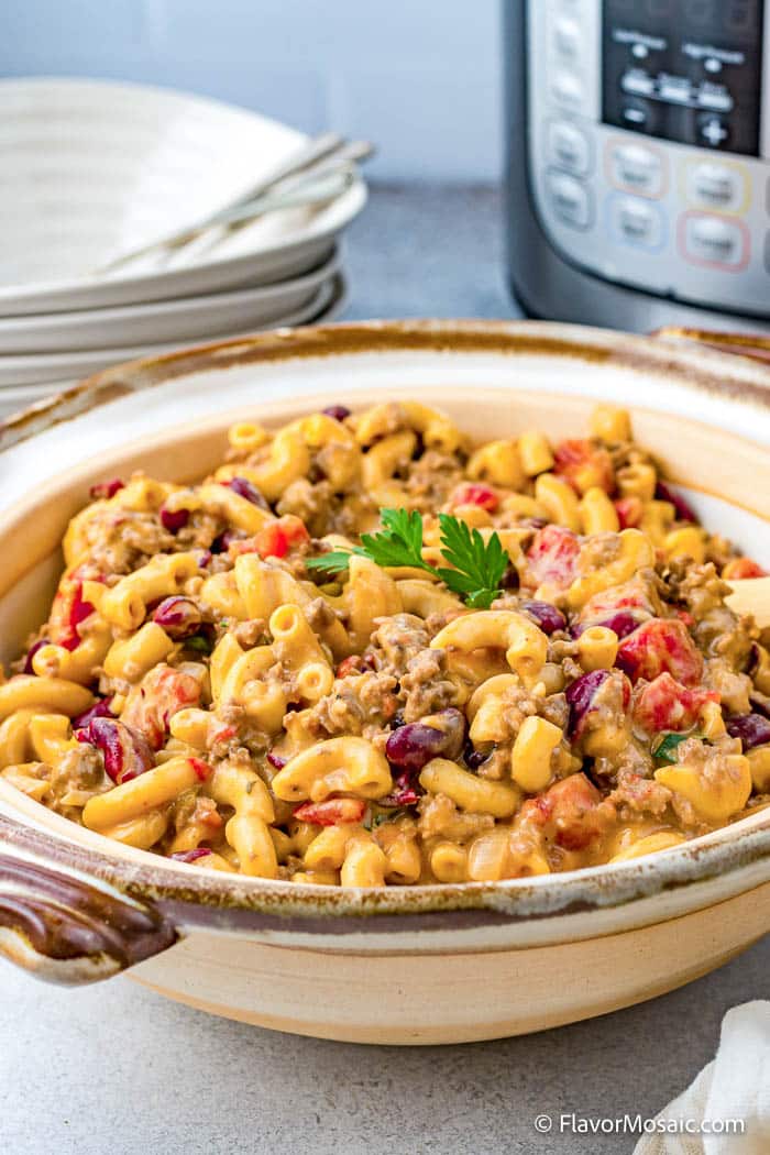 A large serving bowl of Instant Pot Chili Mac with cheesy macaroni, ground beef, and kidney beans in front of a partial view of an Instant Pot and a stack of bowls with spoons in the background.