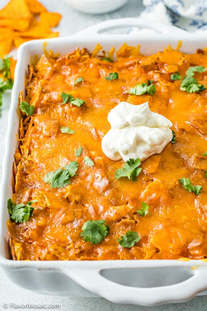 A Chicken Dorito Casserole in a white casserole dish topped with a dollop of sour cream and chopped cilantro leaves over the top.