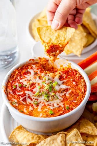 Overhead view of a white bowl with the hot Baked Buffalo Chicken Dip with melted cheese on top and someone dipping a tortilla chip into it.