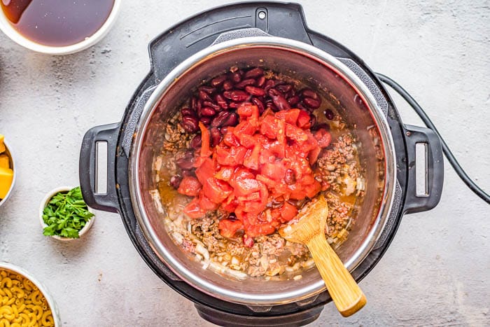 Overhead view of ingredients in the Instant Pot including tomatoes red kidney beans, browned ground beef and onions in broth with a wooden spoon.