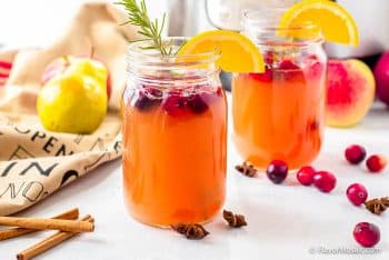 2 glasses of pear apple cider with cranberries, rosemary, and a slice of lemon in each glass, surrounded by cranberries scattered about with pears to the left and an instant pot in the back.