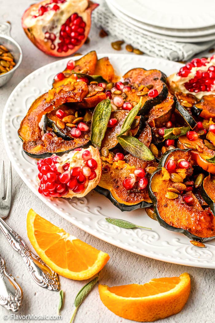 Platter with sliced sweet and spicy roasted acorn squash covered with pomegranate seeds and garnished with a pomegranate that has been sliced open and still has the seeds in it and garnished with sage leaves and an orange slice.