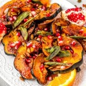 A white platter with slices of seasoned acorn squash topped with pomegranate seeds and sage leaves and garnished with orange slices and a half a pomegranate with the seeds still in it.