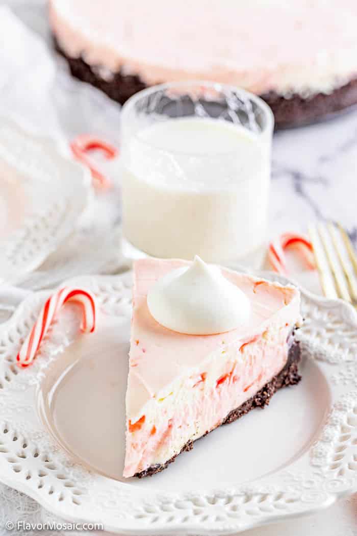 One slice of No Bake Peppermint Cheesecake on a white plate with a small glass of milk and the remainder of the cheesecake in back.