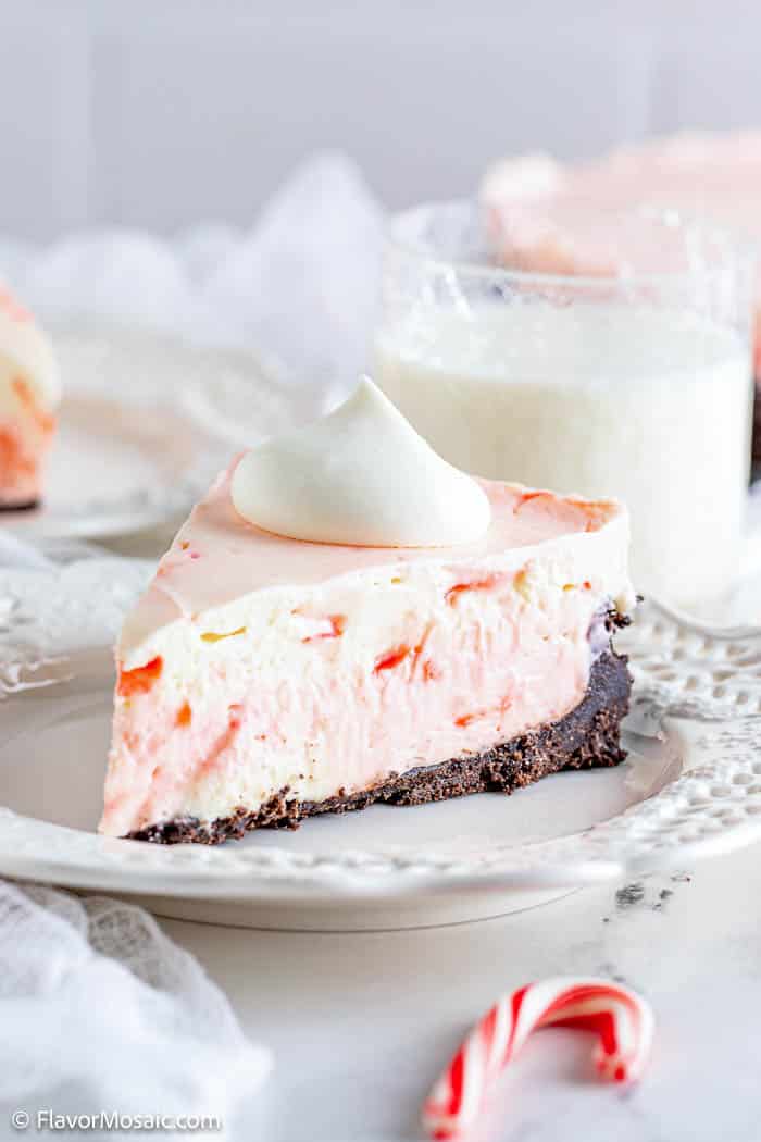 Slice of No Bake Peppermint (Candy Cane) Cheesecake on a white dessert plate with a candy cane next to the plate and a glass of milk in the back.