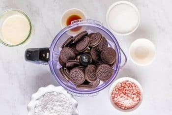Whole Oreos inside a food processor surrounded by individual bowls of other ingredients for the peppermint cheesecake.