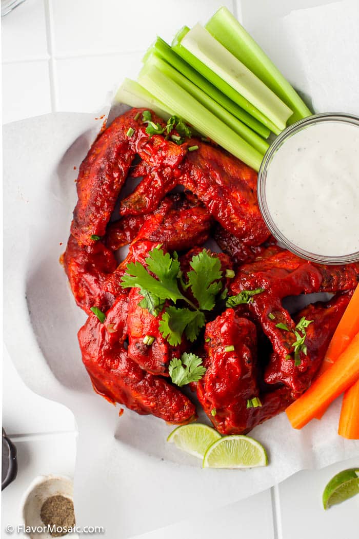 Overhead view of Air Fryer Spicy Honey Chicken Wings in a paper covered bowl with cilantro, celery, and carrot sticks, with 2 lime slices, and small glass bowl of ranch dressing to be used as a dipping sauce.