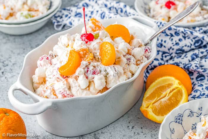 Deep white serving bowl with Ambrosia Salad topped with Mandarin orange slices and a maraschino cherry with a serving spoon at one end of the dish. Sitting on a blue gray background with a white and blue napkin behind it surrounded by orange halves, and partial views of 3 small individual bowls of ambrosia salad. 