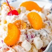 Close up of Ambrosia Salad in a white bowl topped with 3 mandarin oranges and a maraschino cherry with a partial view of a white and blue napkin in the back.