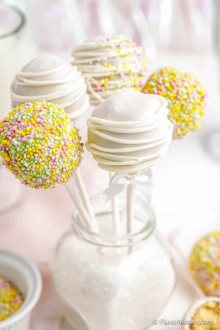 5 decorated white chocolate covered cake pops in a small glass with a white background and small bowls of sprinkles surrounded it at the base.