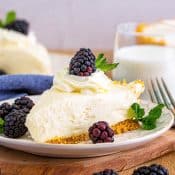 Side view of a slice of plain cheesecake topped with whipped cream and a blueberry on a small white dessert plate sitting on top of wood, surrounded by more blackberries, a blue napkin, and another slice of cheesecake in the back with a small glass of milk in back to the right.