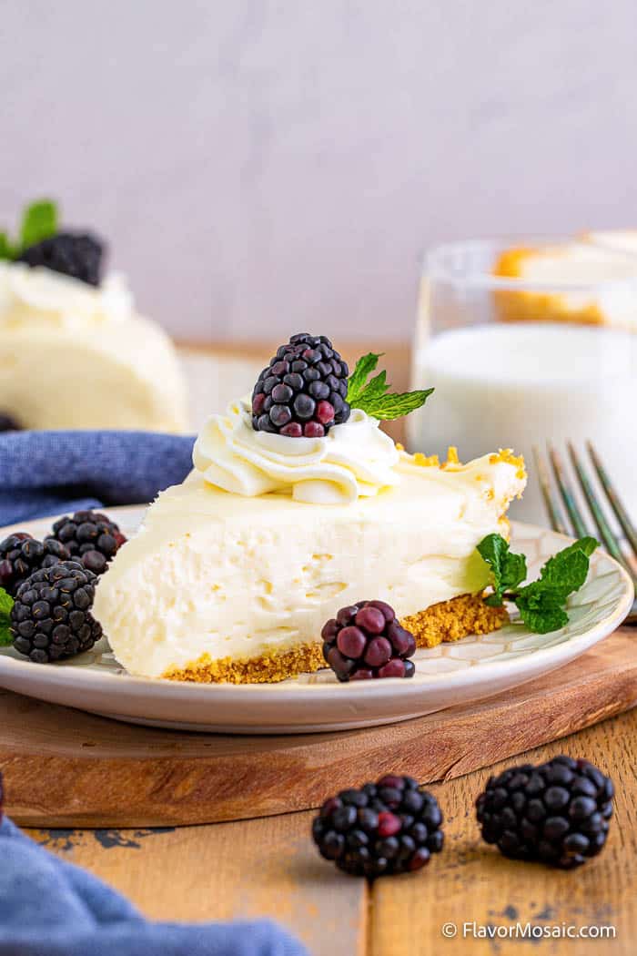 Side view of a slice of plain cheesecake topped with whipped cream and a blueberry on a small white dessert plate sitting on top of wood, surrounded by more blackberries, a blue napkin, and another slice of cheesecake in the back with a small glass of milk in back to the right.
