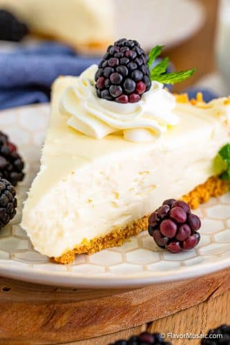 single slice of cheesecake on a white plate on a wood background topped with a swirl of whipped cream and a single blackberry and surrounded by blackberries on the plate.