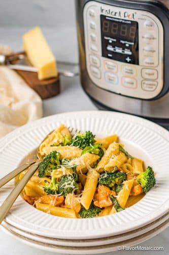 White bowl of chicken cheesy pasta with broccoli in front of an instant pot with a napkin and cutting board with a block of parmesan cheese in the background.