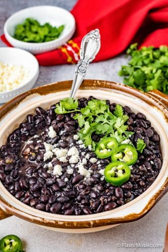Overhead view of a large bowl of black beans topped with chopped onions and sliced jalapeños with a red napkin in the background along with cilantro, a small white bowl of white cheese, and a small bowl of chopped jalapeños.