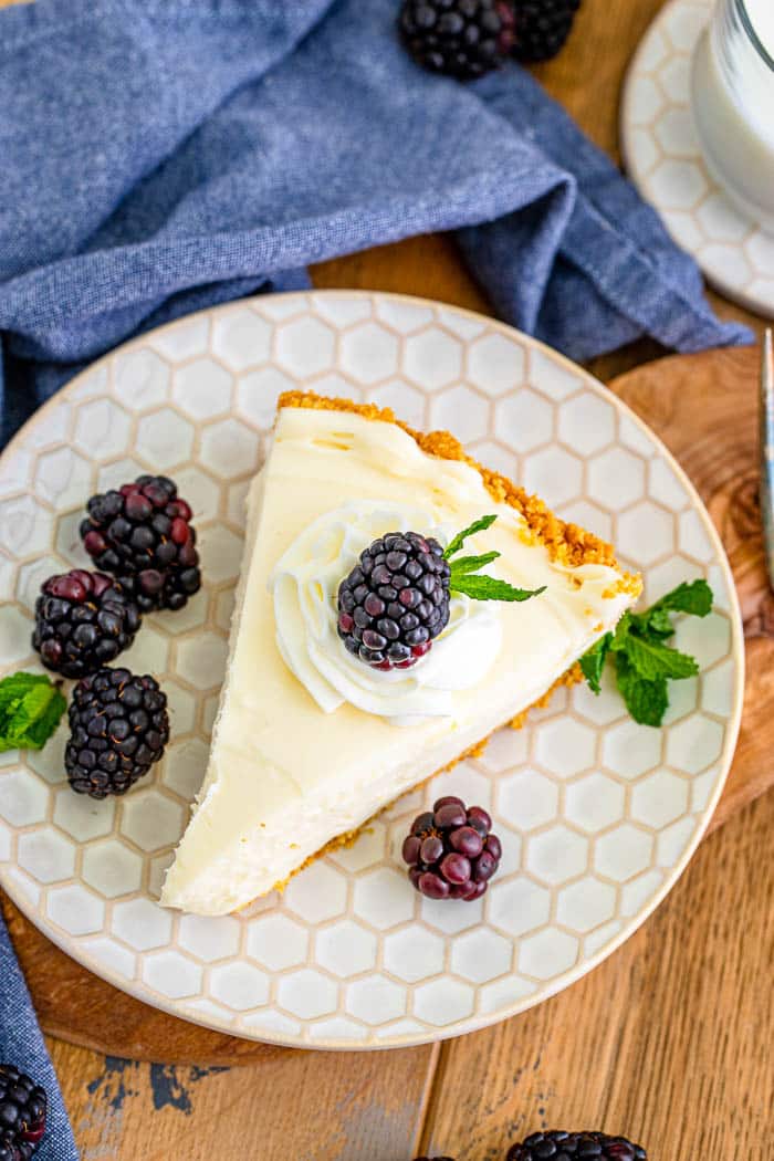 Overhead view of a single slice of cheesecake on a white dessert plate topped with a dollop of whipped cream and a single blackberry, and surrounded by 4 blackberries.