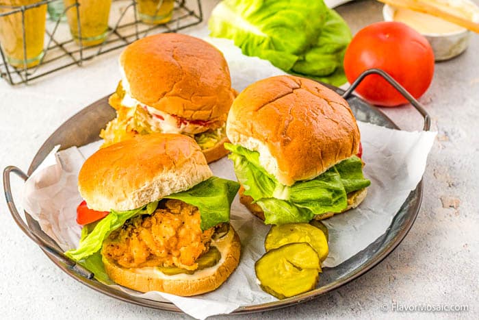 horizontal image showing 3 crispy chicken sandwiches on a metal platter covered with paper, and sliced pickles on the tray as well, with lettuce, tomato and a small bowl of sauce in the background.