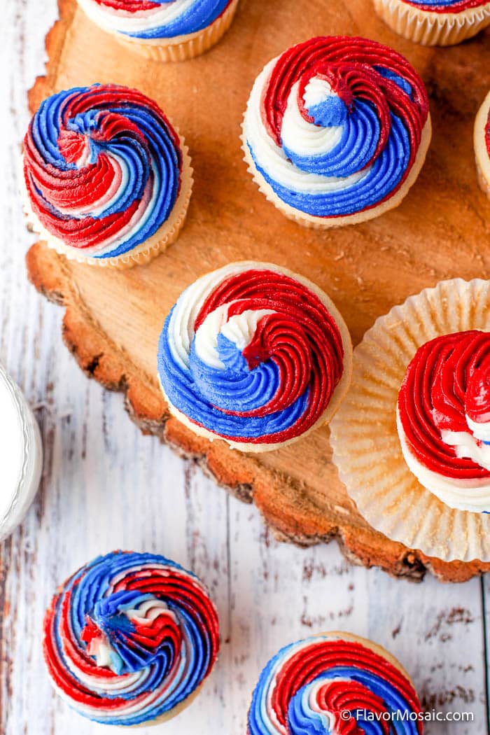 Overhead view of about 6 cupcakes with red white and blue icing, 4 of which are sitting on top of a wood "plate" while 2 others sit below on a rustic white-painted table.