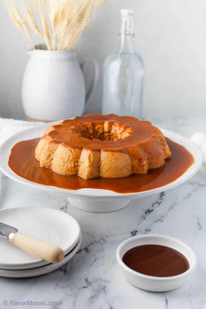 Side view of a whole bundt cake pan shaped flan covered in Mexican caramel (Cajeta) sitting on top of an elevated cake platter.