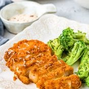 A single air fryer parmesan crusted pork chop on a small white plate with broccoli with a small bowl of a white dipping sauce and a peek of a blue napkin and another plate with an air fryer pork chop.