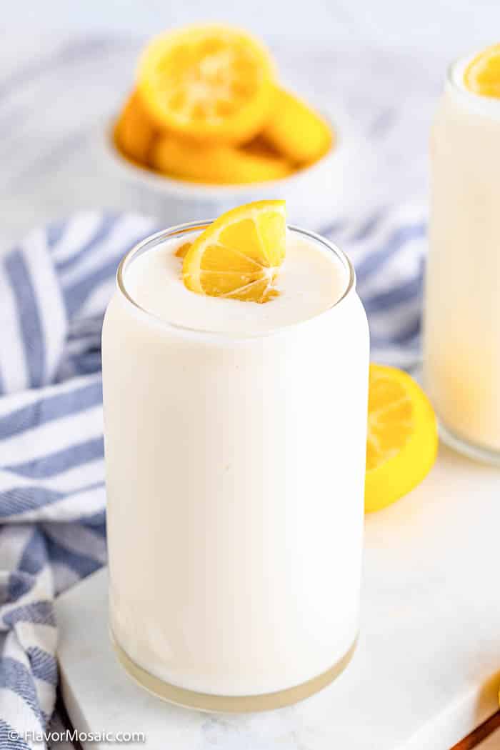 Side view of a glass of Chick Fil A Copycat Frosted Lemonade with a blue and white napkin in the back with a bowl of lemon sliced behind that.