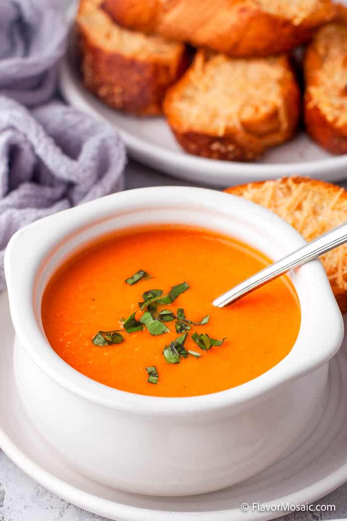 Close up photo of creamy tomato soup with chopped basil in a white soup bowl with a baguette with parmesan cheese in the back and a lavender napkin in the upper left.