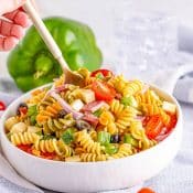 Side view of large white bowl of Tri-Color Rotini Pasta Salad with Italian Dressing with a wooden spoon dipping in to serve it. In the background is a green bell pepper against a white marble background, and cherry tomatoes in the foreground, with the bowl sitting on a light blue-gray napkin.