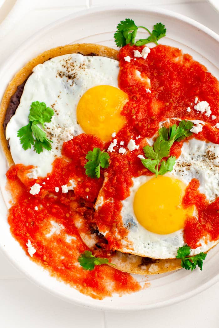 Overhead view of Huevos Rancheros with Huevos Rancheros Sauce and 2 fried eggs garnished with chopped cilantro and served on a white plate.