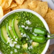 Overhead view of a bowl of salsa verde, green sauce, garnished with avocado slices, chopped onions, serrano peppers, and surrounded by tortilla chips.
