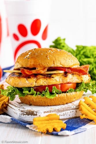 Side view of Chick-Fil-A Grilled Chicken Sandwich with Bacon Cheese and Honey Barbecue Sauce on a rustic white painted board on a blue and white napkin with lettuce in the background.