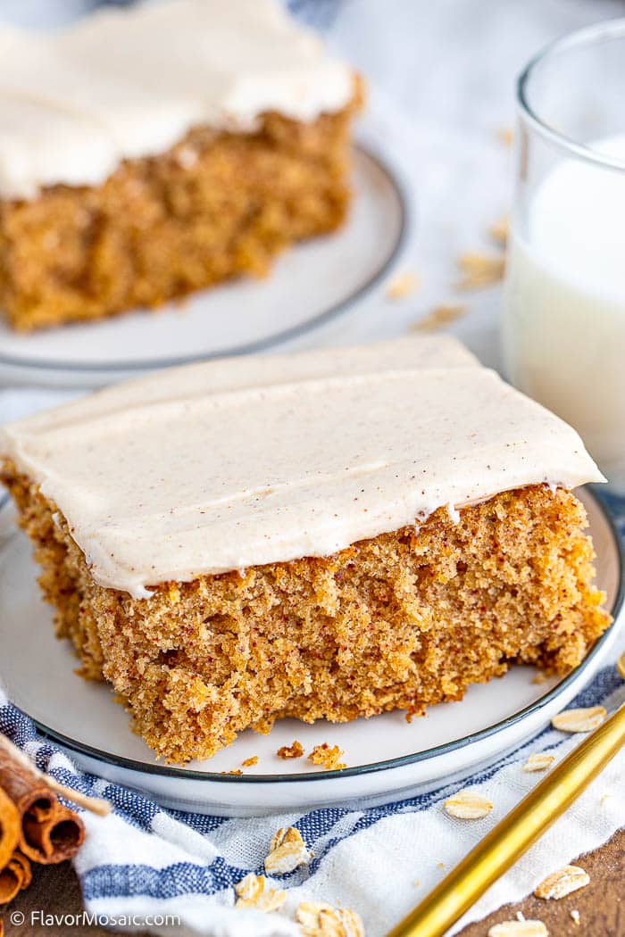 Close up of one serving of oatmeal cake with cream cheese frosting on a white plate with blue trip sitting on a blue and white striped napkin with oats scattered around, the handle of a gold fork in the bottom right, a partial view of cinnamon sticks on the bottom left, a partial view of a glass of milk in the top right and a partial view (blurred) of another piece of cake on a small round white plate with blue trim.