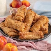 Dark plate with Peach Cobbler Egg Rolls surrounded by peaches and a pink napkin.