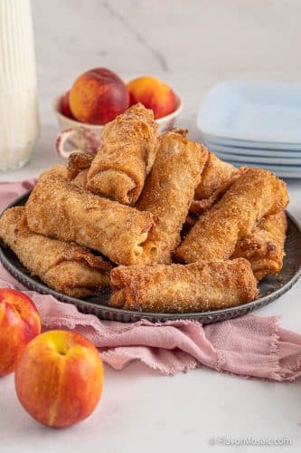 Dark plate with Peach Cobbler Egg Rolls surrounded by peaches and a pink napkin.
