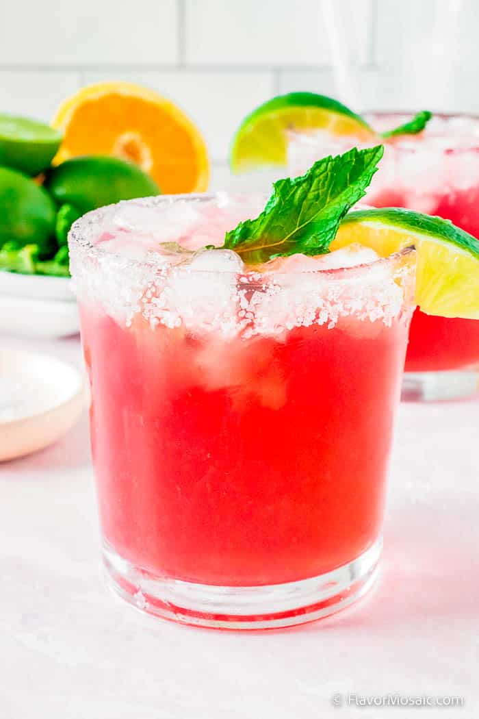 Side view of a salt-rimmed glass with a pink margarita with cranberry juice cocktail, garnished with a mint leaf and slice of lime, with another margarita glass in the background along with a plate of limes and a sliced orange.