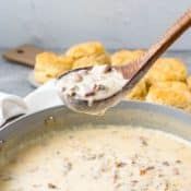 Spoonful of sausage and bacon gravy held above a skillet of gravy with biscuits in the background.