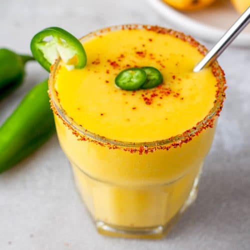 Side overhead view of a single glass of mango jalapeno margarita with Tajin salt on the rim of the glass and garnished with jalapeno slices and Tajin seasoning with a stirrer in it. Jalapeno slices in the bottom left, 2 jalapenos in the middle left, and mangos in the top right all on a gray background.