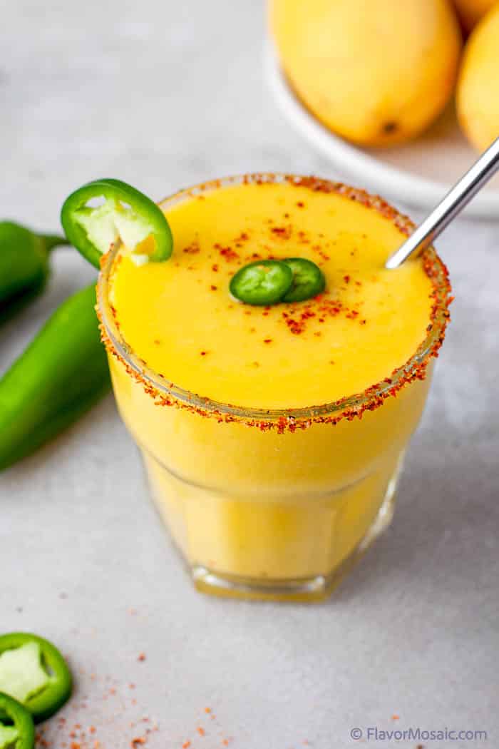 A close up view of a single mango jalapeno margarita with tajin salt on the rim a jalapeno slice on the side and a garnish of small jalapeno slices.