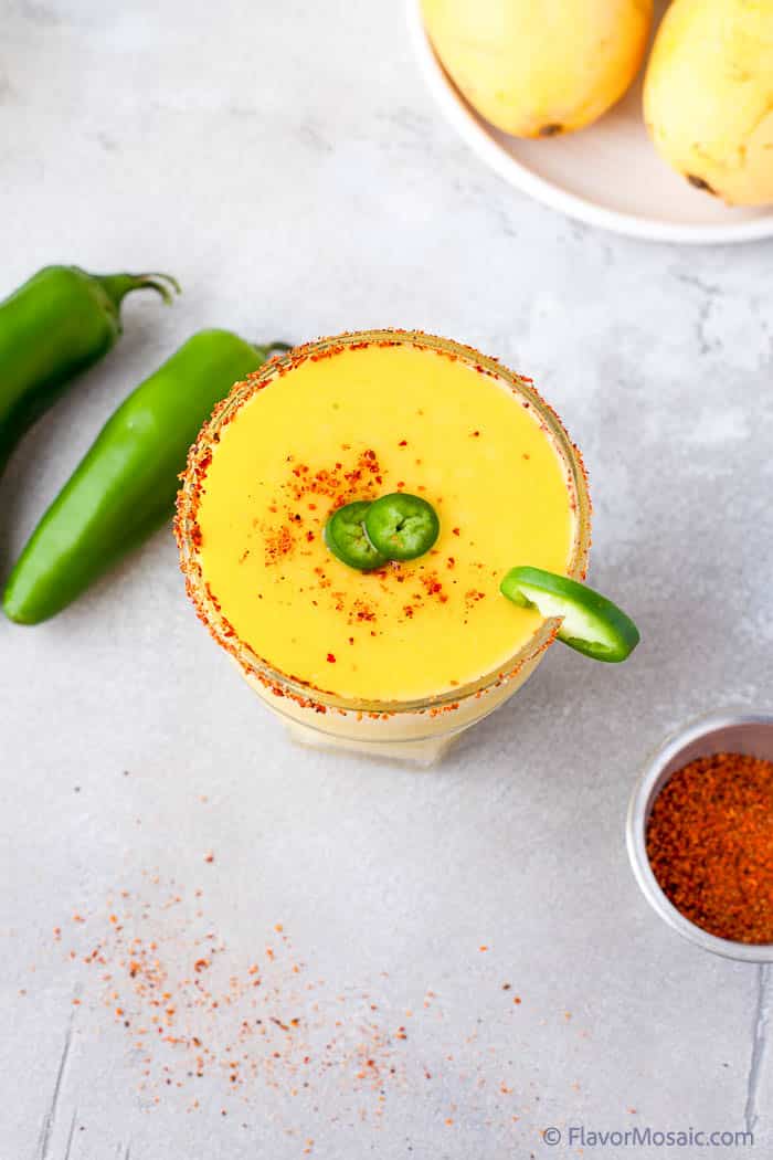 An overhead view of a single mango jalapeno margarita garnished with tajin and jalapeno slices, and a jalapeno slice on the rim of the glass. Jalapenos on the left and a small bowl of tajin on the bottom right.