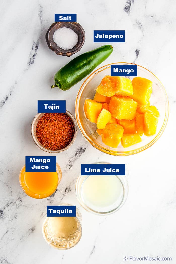 An overhead photo of the labeled ingredients for a mango jalapeno margarita, each ingredient in separate bowls, except the jalapeno, on a white marble background.