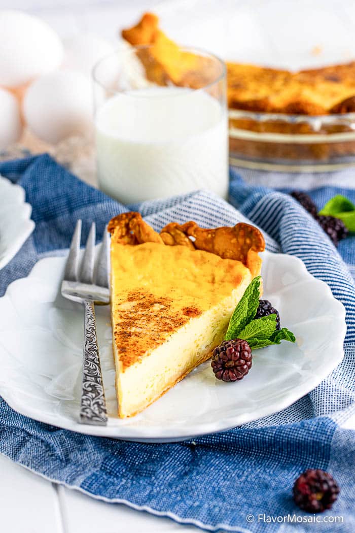 A slice of custard pie on a white plate with a metal fork on the left and 2 blackberries and mint leaves on the right, all sitting on a denim blue and white napkin with a glass of milk and eggs in the background.