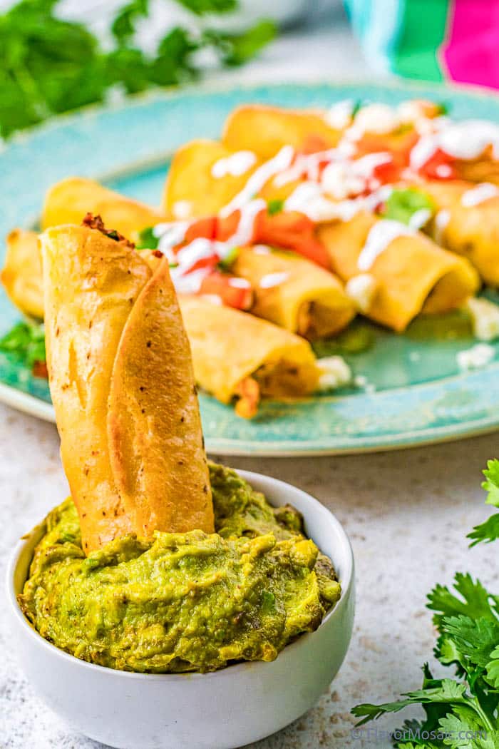 A Chicken and Cream Cheese Taquito standing straight up in a small bowl of guacamole, with the rest of the plate of taquitos in the background on an aqua plate.