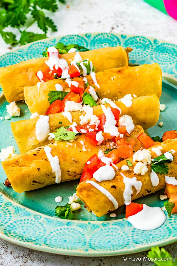 5 chicken and cream cheese taquitos on an aqua plate garnished with sour cream, chopped tomatoes, chopped cilantro and Mexican queso fresco.