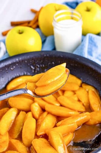Fried apples in a skillet and a spoon holding a spoonful of fried apple slices above the skillet with 3 golden apples with a glass of milk and blue napkin in the background.