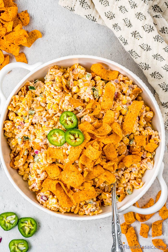 Overhead view of a white bowl with Frito Crack Corn Salad topped with Frito corn chips, surrounded by sliced jalapeños, corn chips, and a napkin.