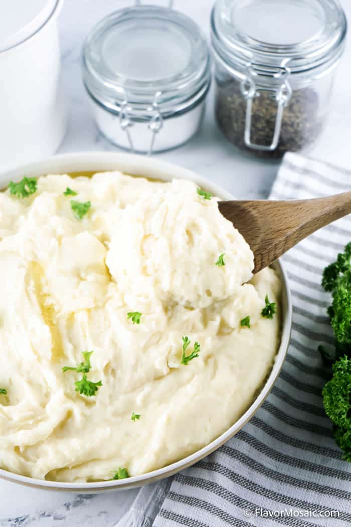 Overhead view of mashed potatoes with chopped parsley with a wooden spoon starting to lift mashed potatoes out of the bowl. A blue-gray and white stripped napkin in the background. With small glass mason jars of salt and pepper in the back.